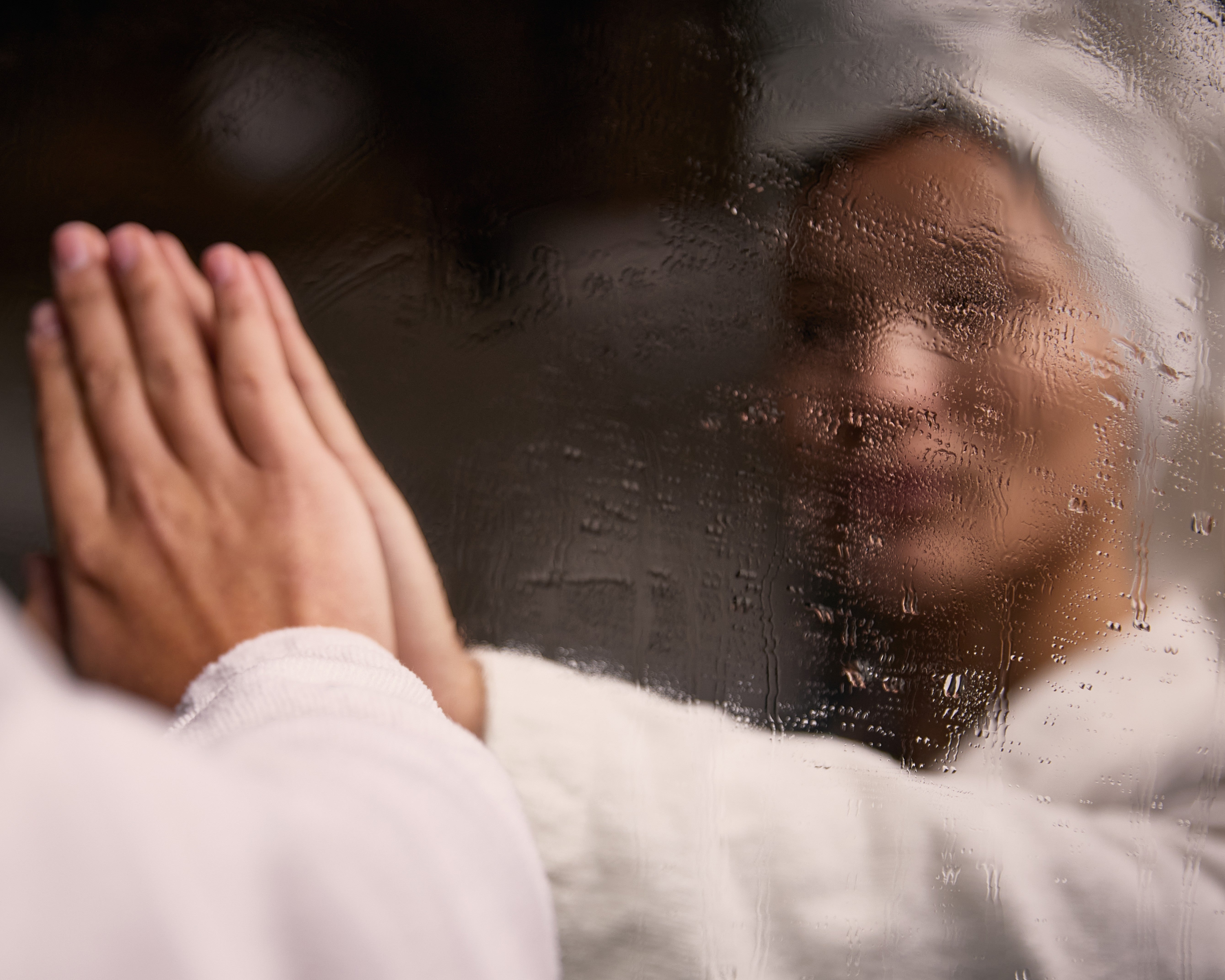 hands pressed against a rain-covered window, face softly blurred behind the glass