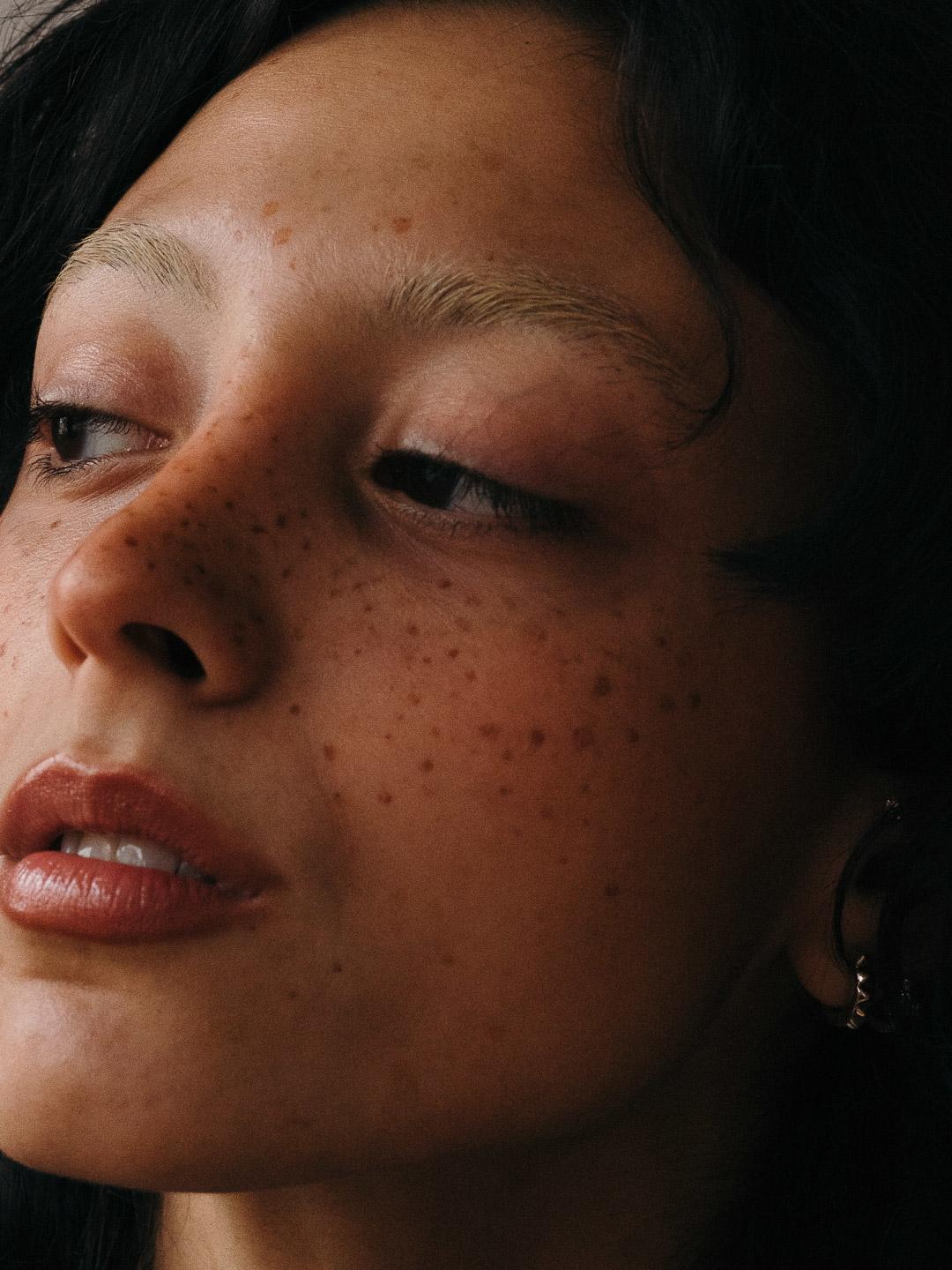 Dark hair, freckles, looking away A close-up portrait of a young woman with dark hair and freckles, eyes downcast, expression thoughtful and inward — a quiet moment of self-reflection.