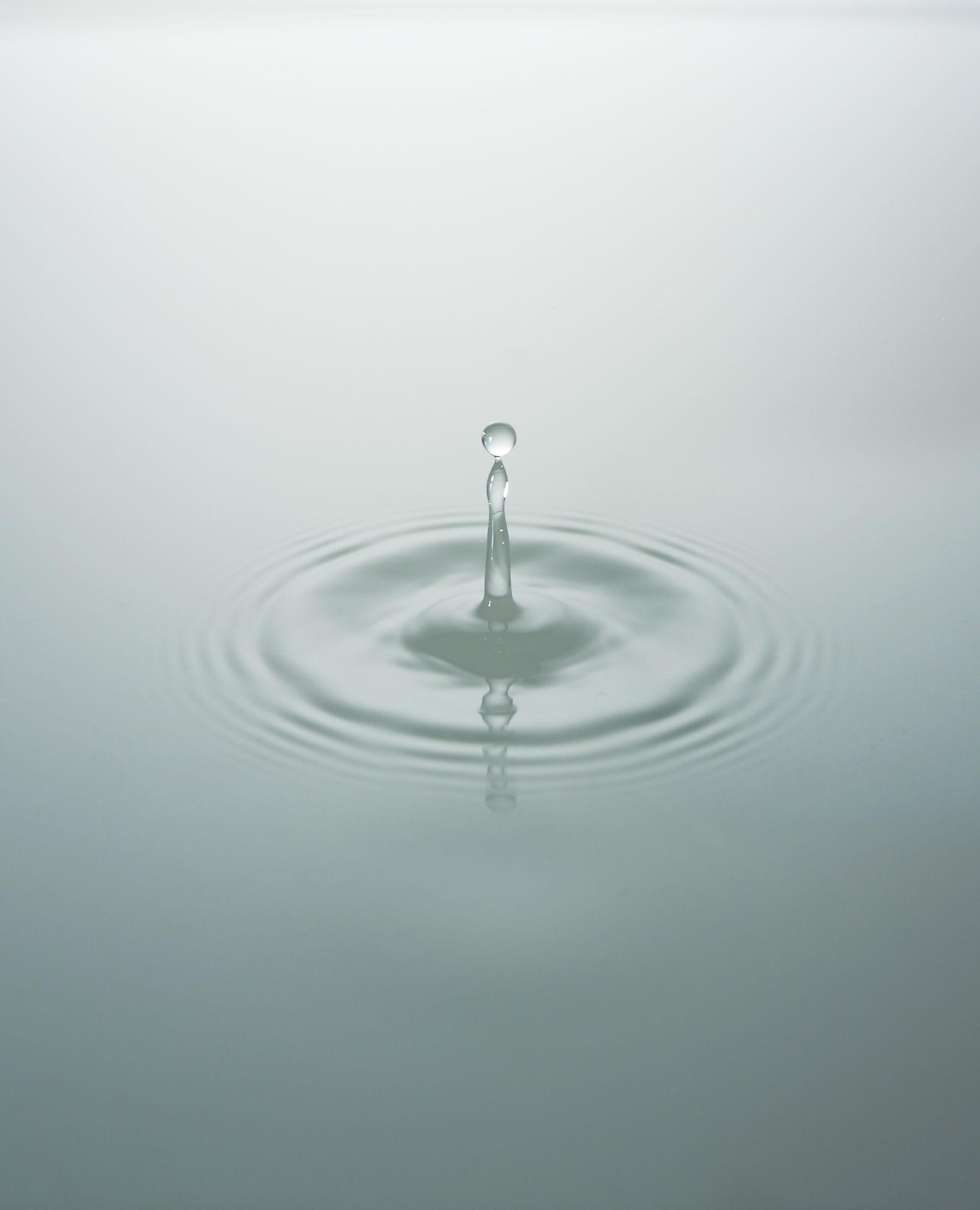 Water drop A single water droplet caught mid-rise above a still, pale surface, ripples spreading outward in perfect circles — a moment of quiet impact and gentle change.