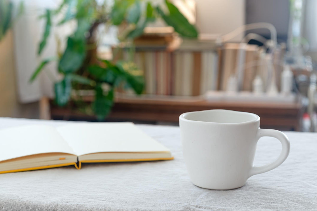 A white ceramic mug and an open journal resting on a linen tablecloth, with soft natural light and blurred greenery in the background.